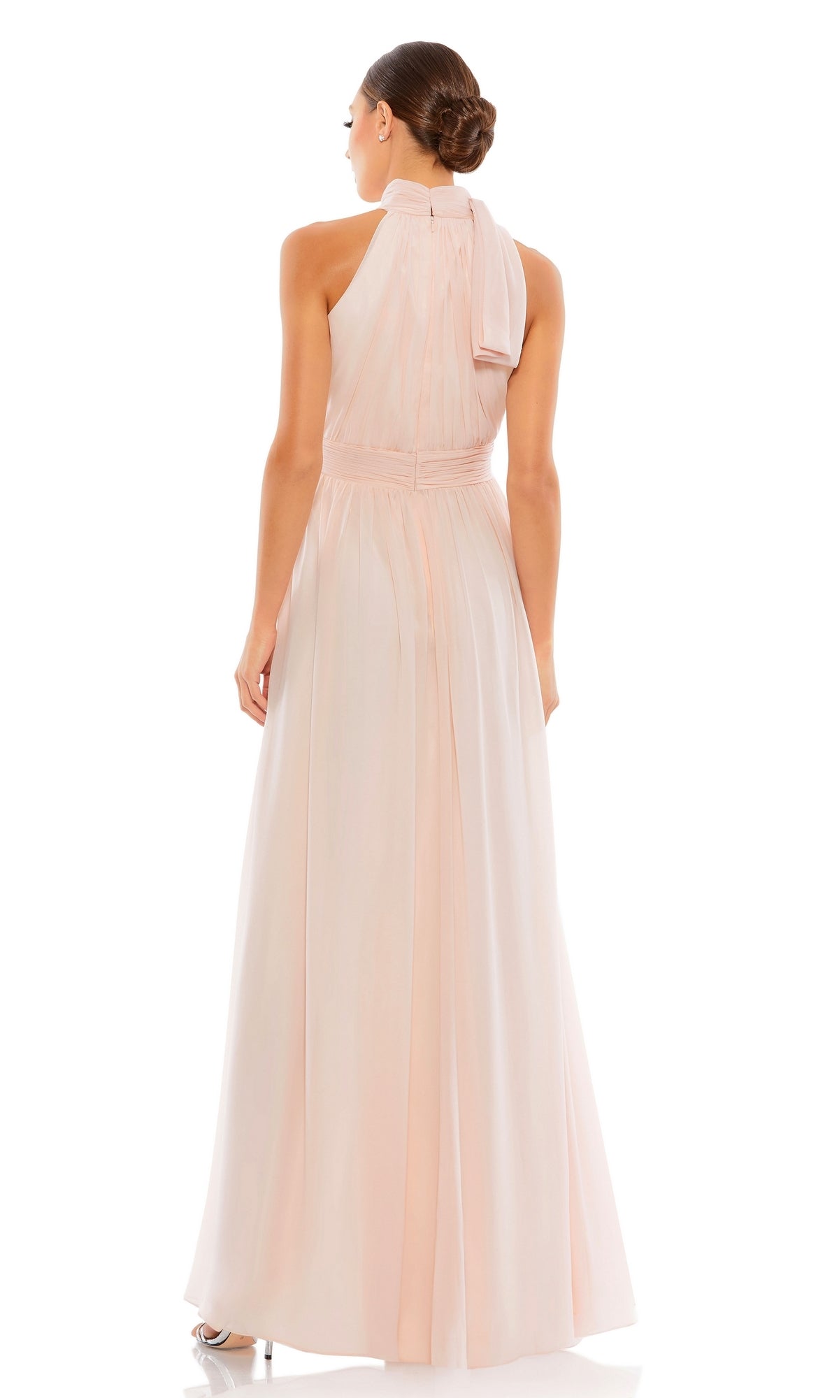A woman with her hair in a neat bun stands facing away, wearing the Mac Duggal 55035 high-neck, floor-length formal dress in pale pink with a bow at the neck.